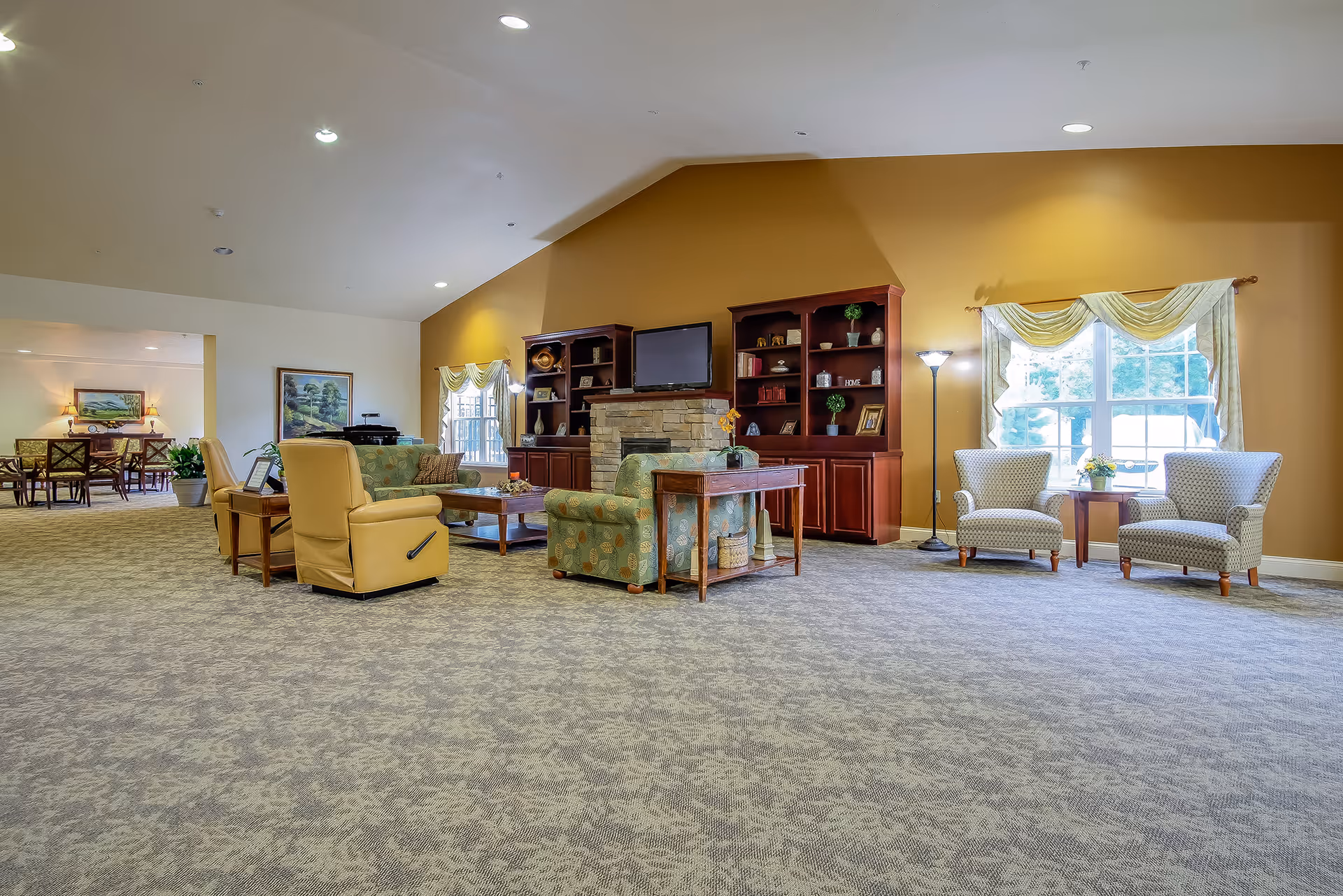 A spacious living room area in an assisted living facility with beige carpet and a high ceiling. The room features a stone fireplace with a TV mounted above it, flanked by two wooden bookshelves filled with decorative items. There are several upholstered chairs and sofas arranged around a wooden coffee table. Two large windows with draped curtains allow natural light into the room. In the background, a dining area with tables and chairs is visible.