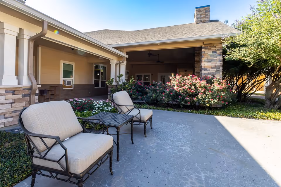 Outdoor patio area at Bristol Park at Cleburne Assisted Living & Memory Care featuring two cushioned metal chairs and a small metal table on a concrete surface, surrounded by flowering bushes and greenery, with part of the building visible in the background.