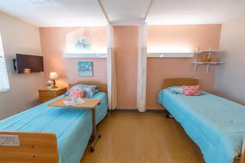 A bright and clean room with two single beds separated by a privacy curtain. Each bed has a turquoise bedspread and a decorative pillow. There is a small wooden nightstand with a lamp and a mounted TV on the left wall. The walls are painted in soft peach and white tones, and there are two small paintings above the beds.