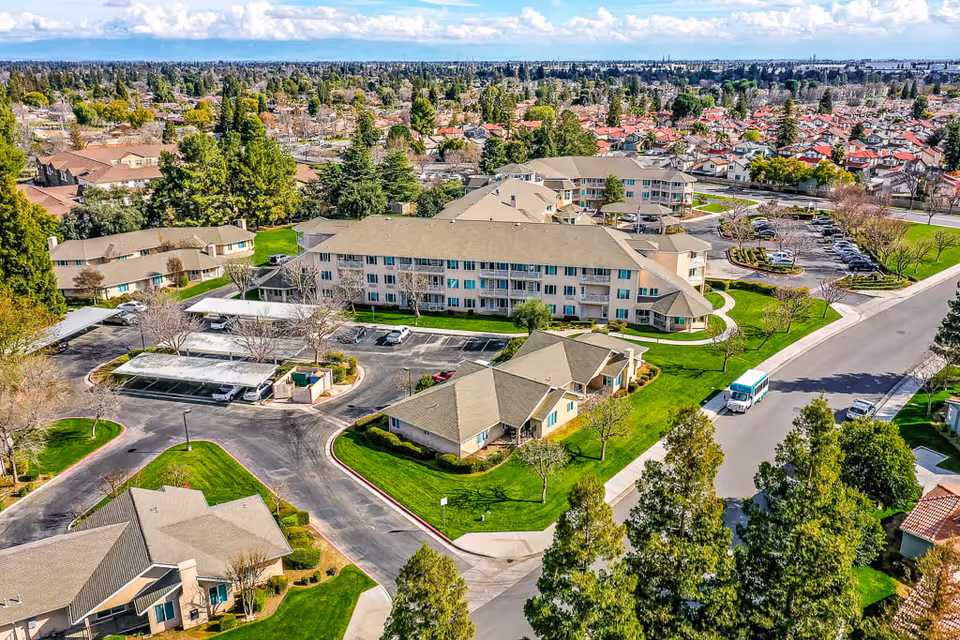 Aerial view of Solstice Senior Living at Bakersfield showing multiple residential buildings surrounded by green lawns, trees, parking areas, and roads with a bus and cars. The neighborhood and cityscape extend into the distance under a partly cloudy sky.