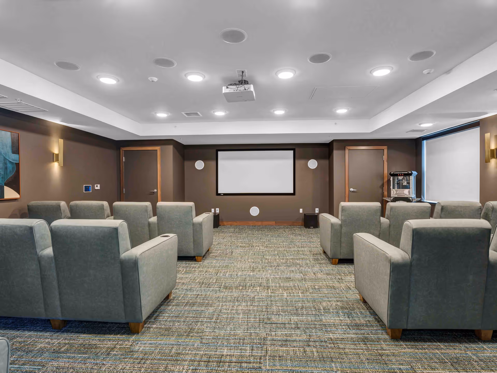 A senior living facility media room with multiple rows of gray upholstered armchairs facing a large blank projector screen on a brown wall. The room has recessed ceiling lights, two closed wooden doors on either side of the screen, a popcorn machine near the right door, and a patterned carpeted floor.
