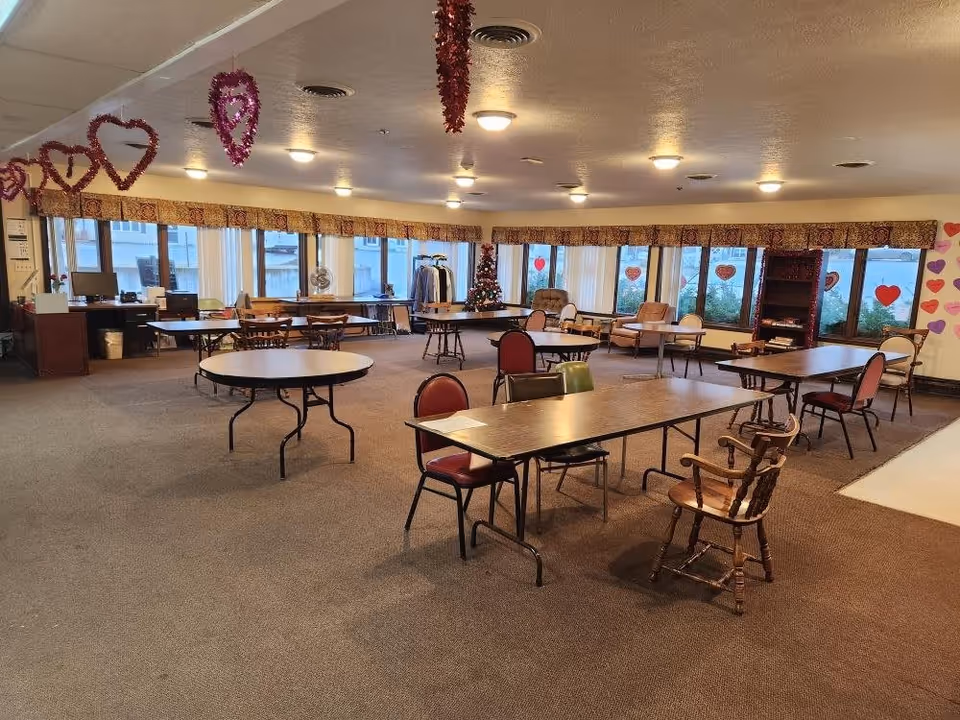 A spacious common room in Glenwood Assisted Living decorated with hanging heart-shaped garlands and Valentine-themed window decorations. The room contains multiple tables and chairs arranged for group seating, with large windows letting in natural light. There is a Christmas tree in the corner and a few armchairs near the windows.