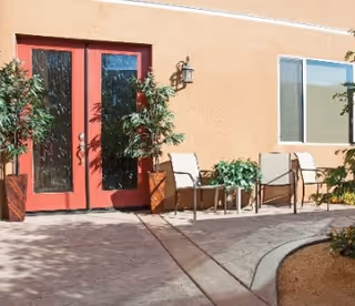 Outdoor courtyard with red double doors, patio chairs, potted plants, and a curved walkway against a peach-colored building.