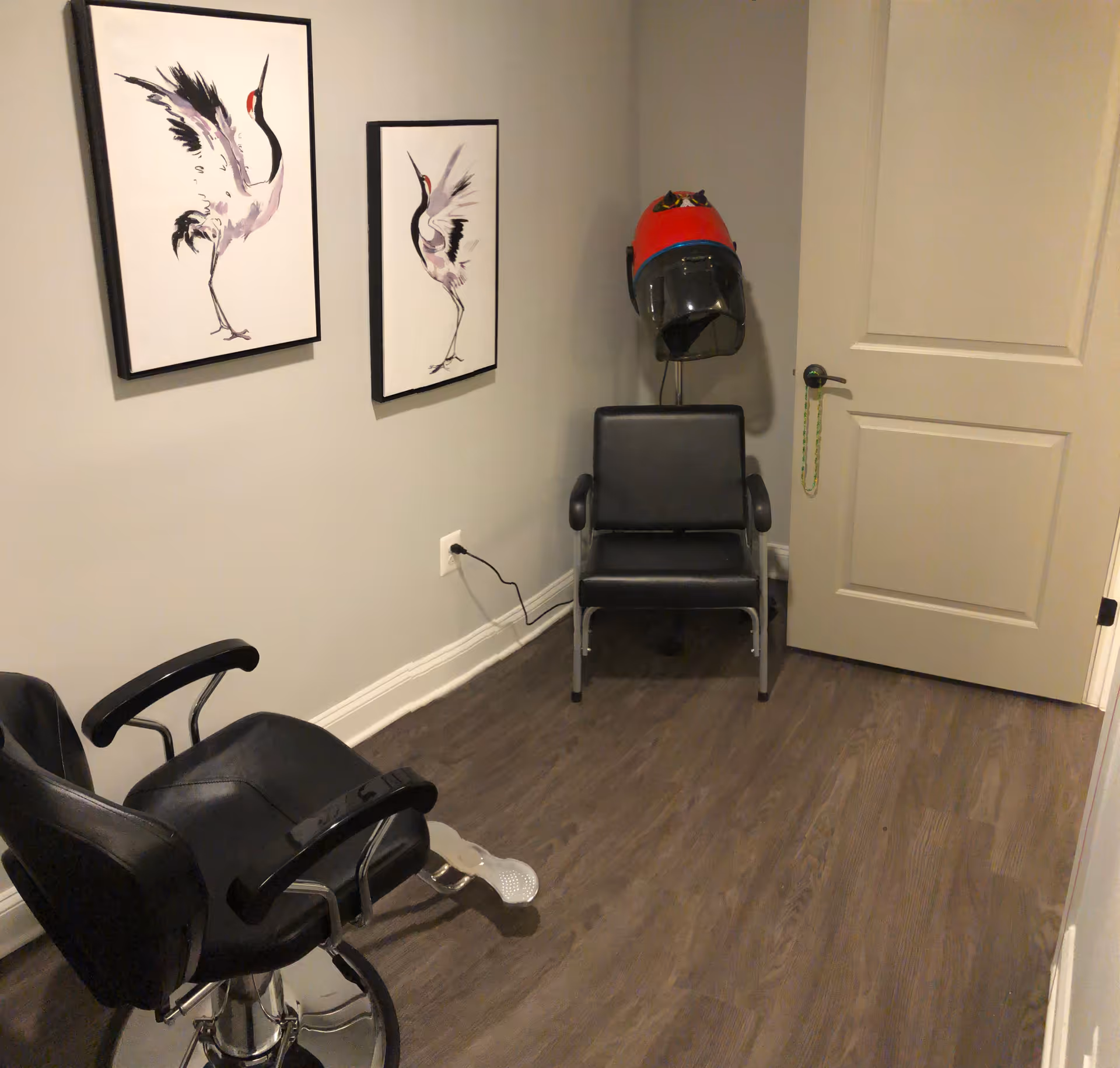 A small room with two black salon chairs, one with a footrest and the other positioned under a red hair dryer hood. The walls are light-colored and decorated with two framed pictures of cranes. The floor is wood, and there is a closed beige door with a green beaded chain hanging from the handle.