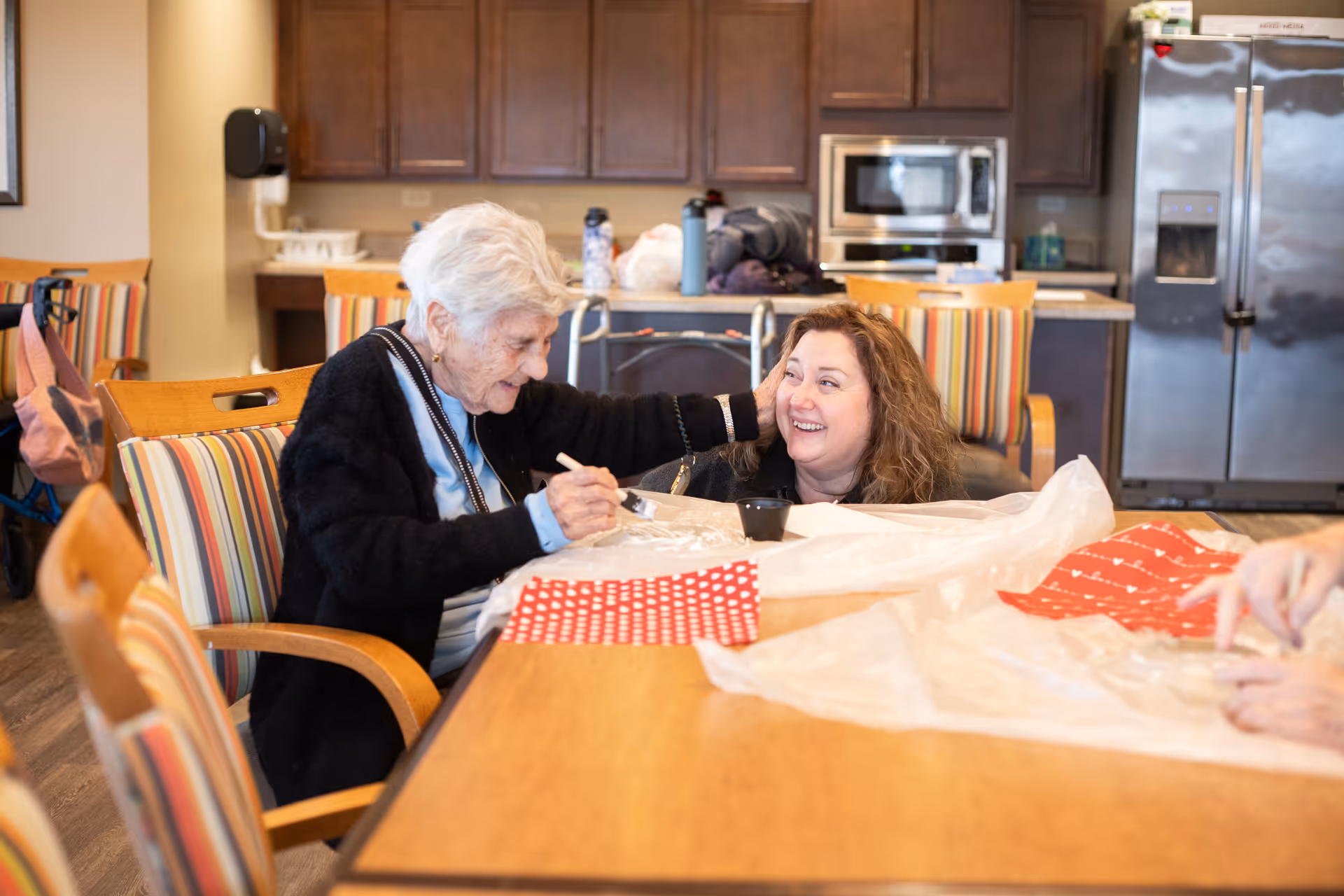 An older woman and another woman smile while working on crafts at a table in a communal dining area with a kitchen in the background.