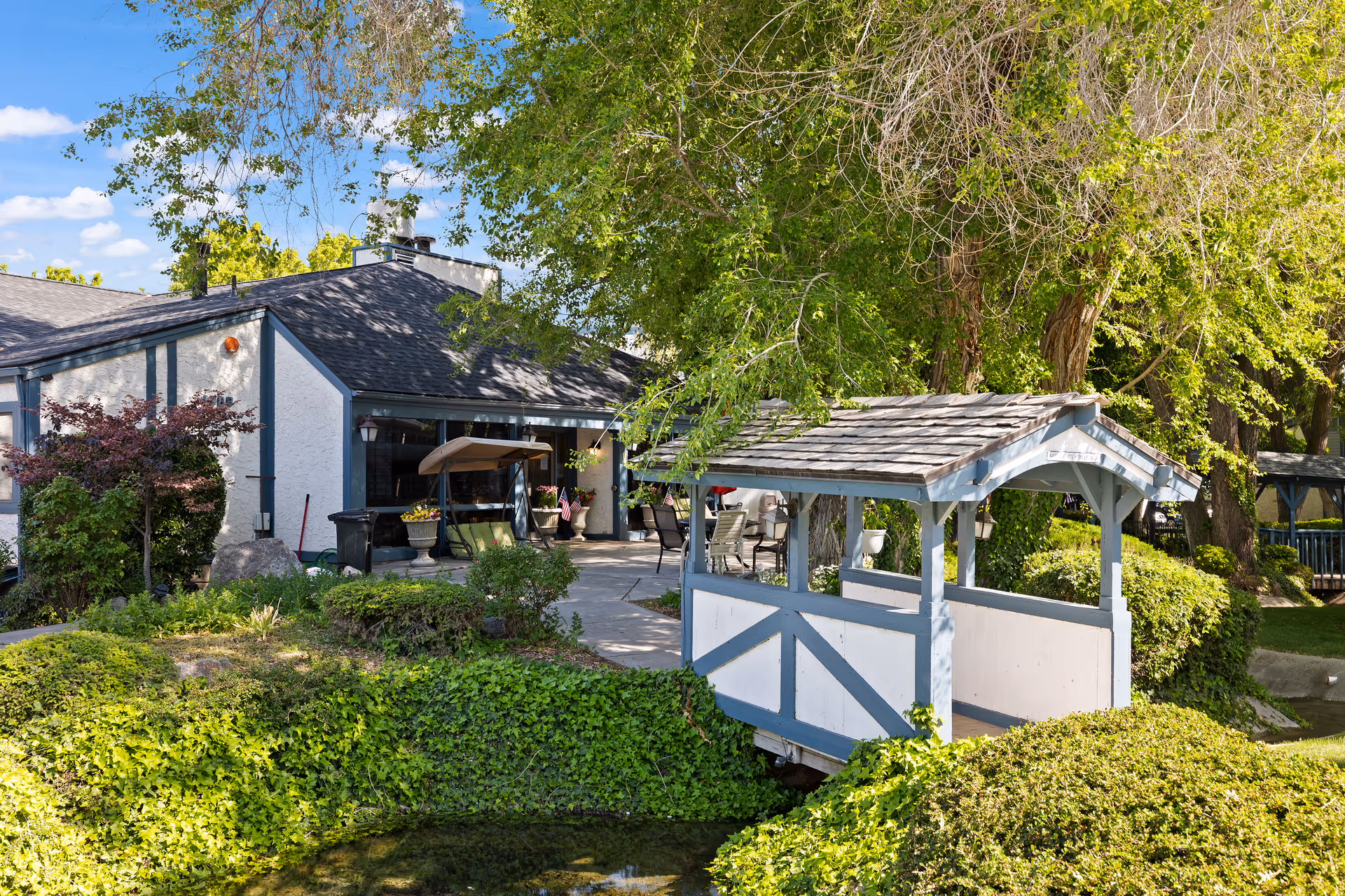 A peaceful outdoor scene at Cove Point Retirement featuring a small covered wooden bridge over a pond surrounded by lush greenery and trees. In the background, there is a patio area with outdoor furniture adjacent to a building with a dark roof and light-colored walls.