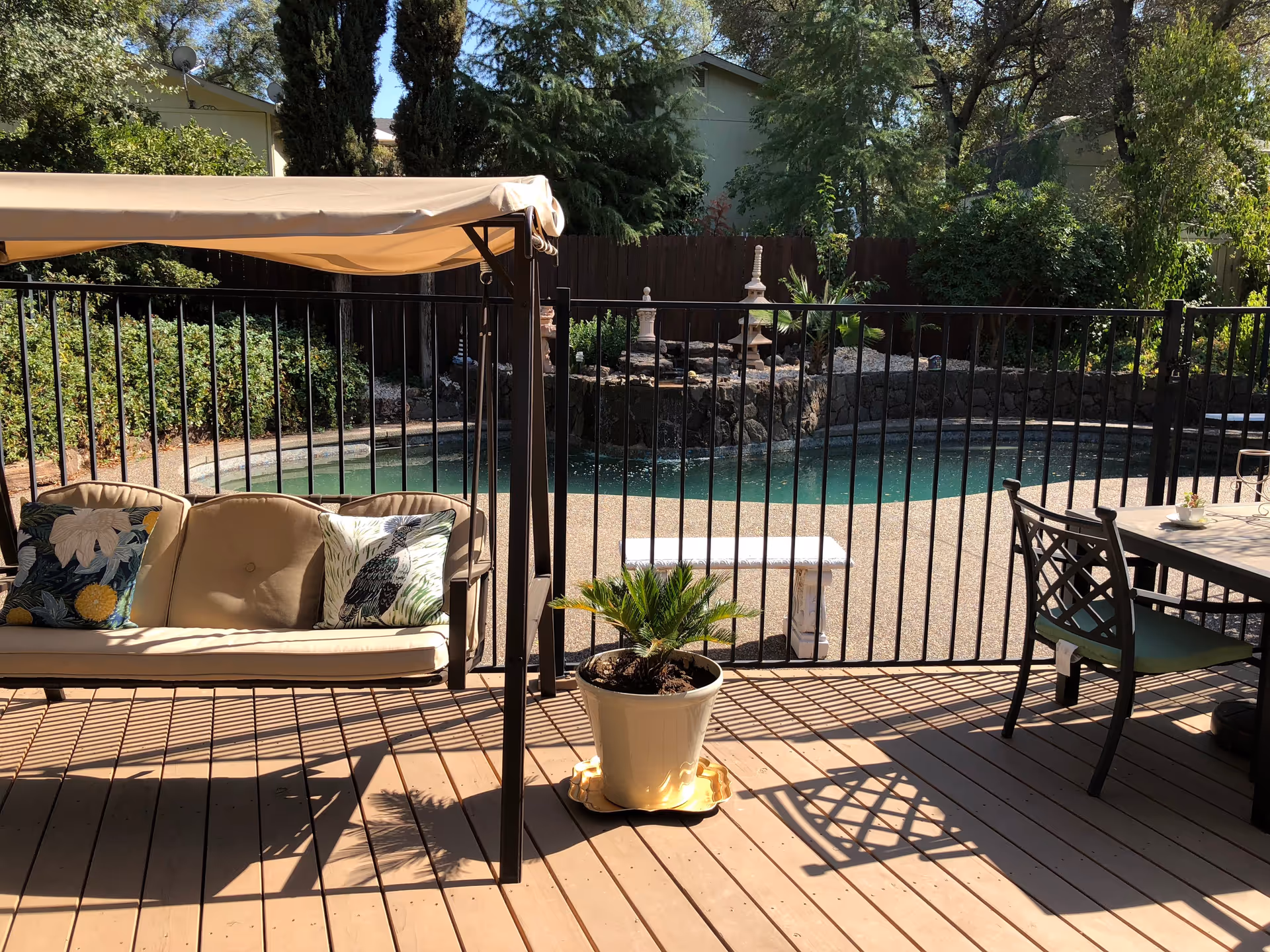 Outdoor patio area with a cushioned swing featuring two decorative pillows, a potted plant on a wooden deck, a metal fence enclosing a small pool with a stone border, and a table with chairs on the right side.
