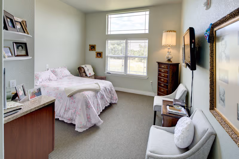 A cozy bedroom in an assisted living facility with a single bed covered in floral bedding, a brown armchair next to the window, a tall wooden dresser with a lamp, two upholstered chairs with a small table between them, framed photos on the walls and shelves, and a flat-screen TV mounted on the wall.