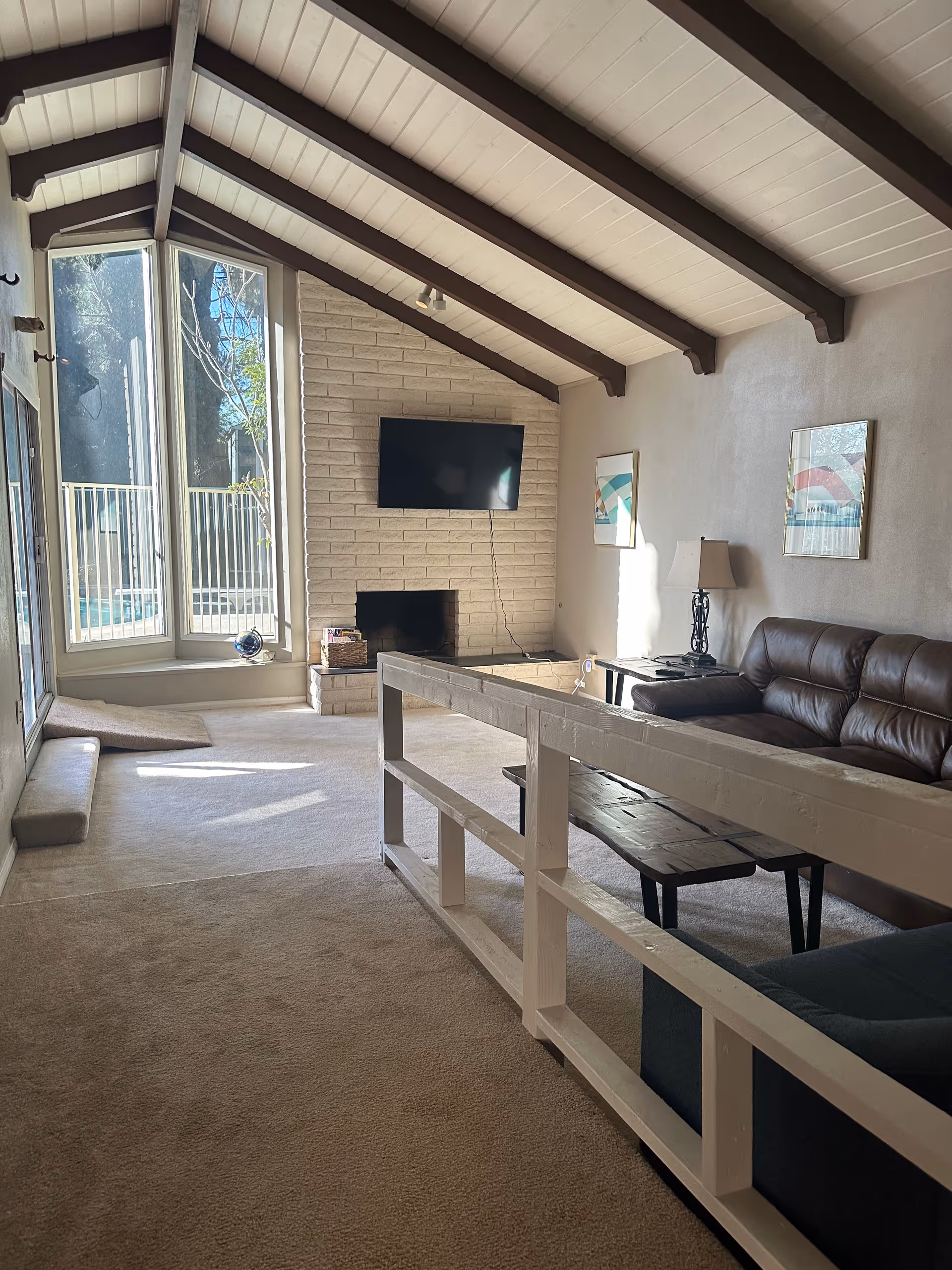 A bright living room with a high vaulted ceiling featuring dark wooden beams and white paneling. There is a large window with a view of an outdoor area, a brick fireplace with a mounted flat-screen TV above it, a brown leather sofa, a wooden coffee table, a side table with a lamp, and framed artwork on the wall. The room has beige carpeting and a white wooden railing separating the seating area.