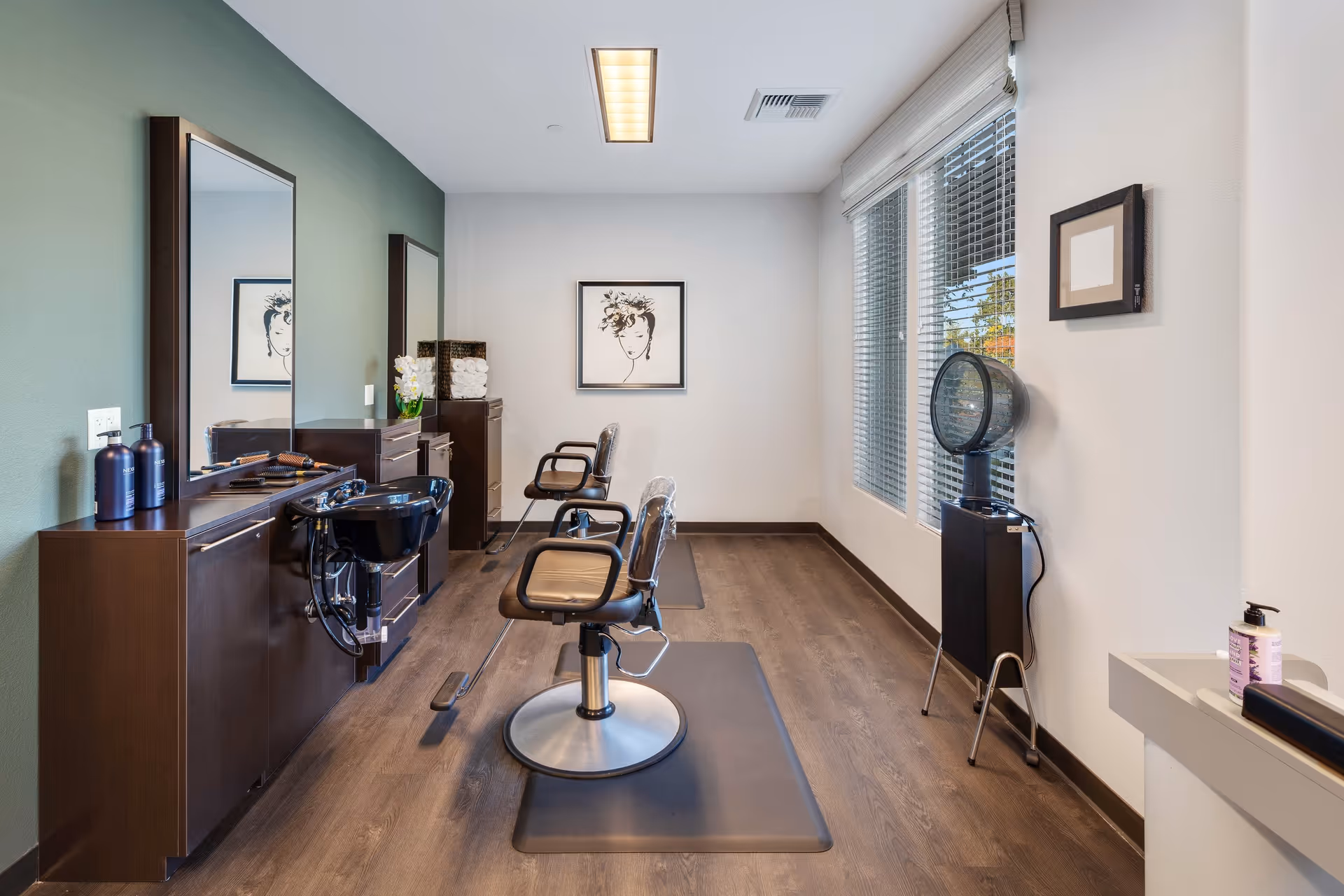 Interior view of a hair salon area in a senior living facility with two salon chairs on mats, a black wash basin, large mirrors, hair care products on a wooden counter, a hair dryer, and large windows with blinds letting in natural light.