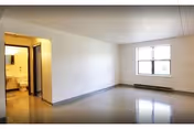 Empty room with a large window letting in natural light, a door leading to a bathroom with a visible toilet and sink, and plain white walls with a gray baseboard.