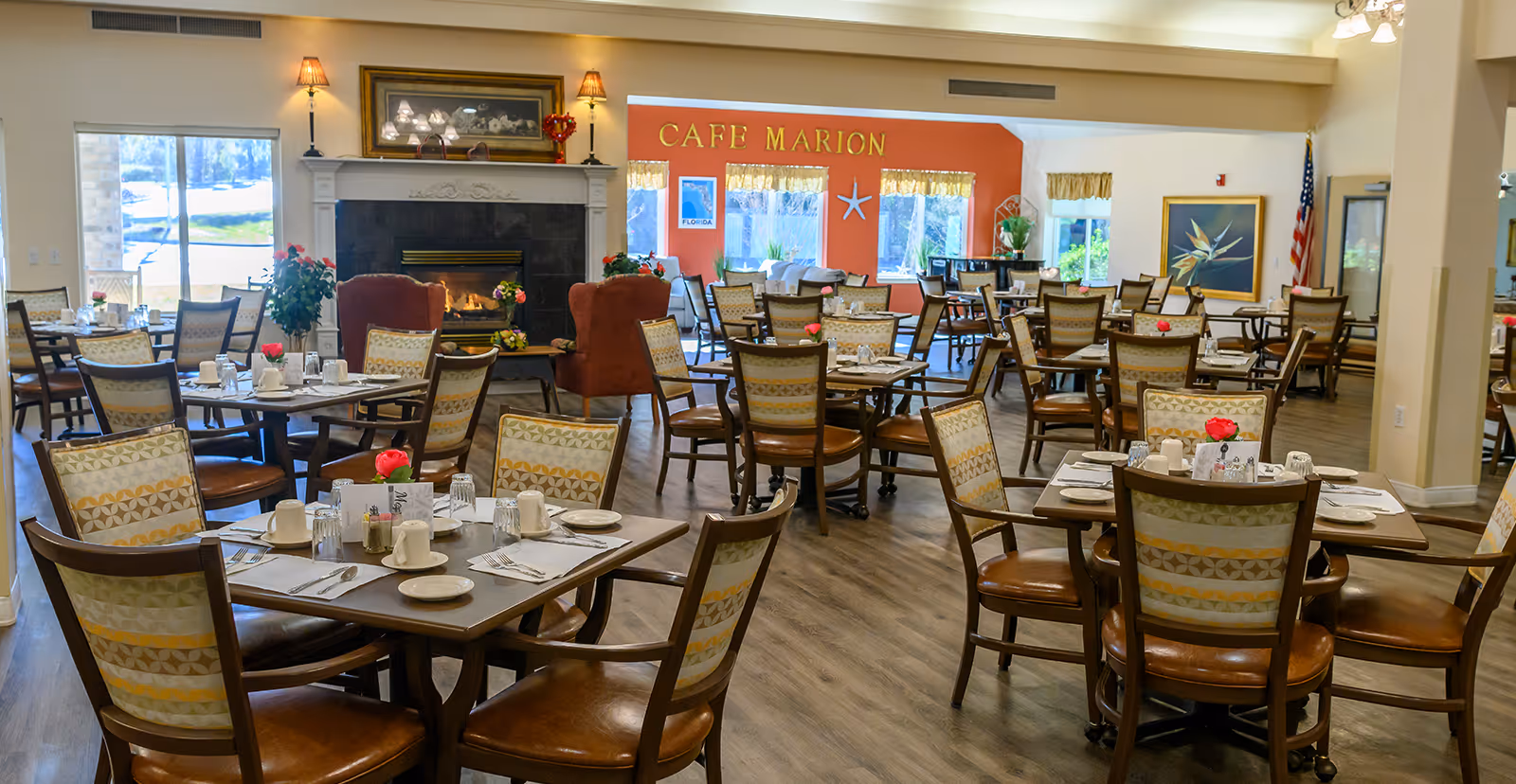A spacious dining room with multiple tables and chairs arranged neatly. Each table is set with white cups, plates, utensils, and a small vase with a red flower. There is a fireplace with a lit fire and two armchairs in front of it. The back wall is painted orange with the words 'CAFE MARION' displayed above three windows with yellow curtains. The room has wooden flooring and is well-lit with natural light coming through the windows.