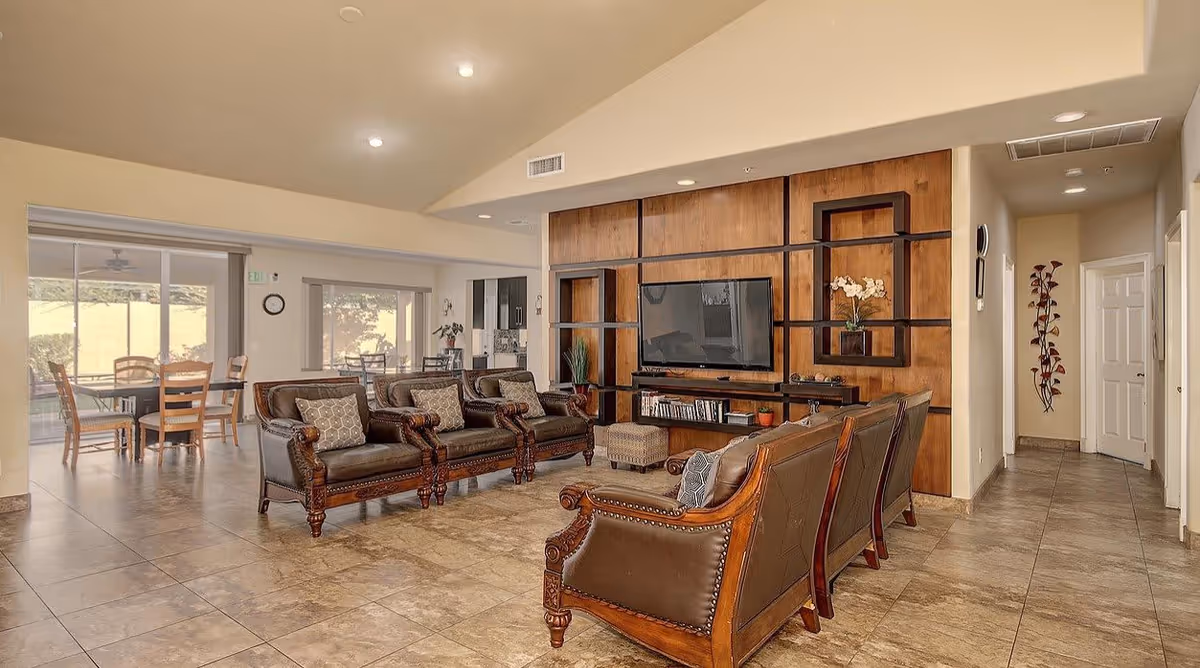 A spacious living room area with a set of brown leather armchairs and sofas arranged around a wooden entertainment wall unit with a flat-screen TV. The room has tiled floors, recessed lighting, and large windows allowing natural light to enter. In the background, there is a dining area with tables and chairs near sliding glass doors leading outside.