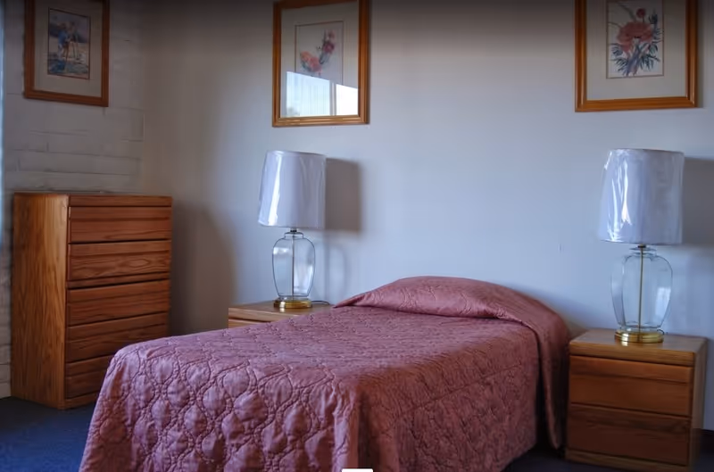 A simple bedroom with a single bed covered in a pink quilted bedspread. On each side of the bed is a wooden nightstand with a glass lamp that has a white lampshade. There is a wooden chest of drawers to the left of the bed. Three framed floral pictures hang on the wall above the bed.