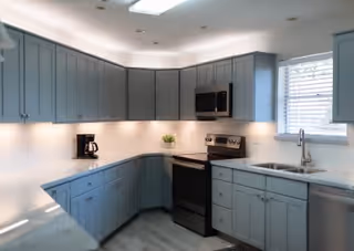Light-filled kitchen with pale blue cabinets, black stove and microwave, and a sink beneath a window.
