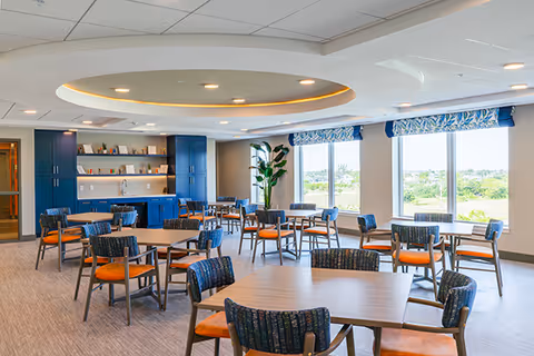 A bright and spacious dining room with multiple wooden tables and chairs featuring blue patterned backs and orange cushions. Large windows with blue patterned valances allow natural light to fill the room, and a built-in blue cabinet with a countertop and sink is visible along one wall. The ceiling has a recessed circular light fixture, and a tall green plant is placed near the windows.