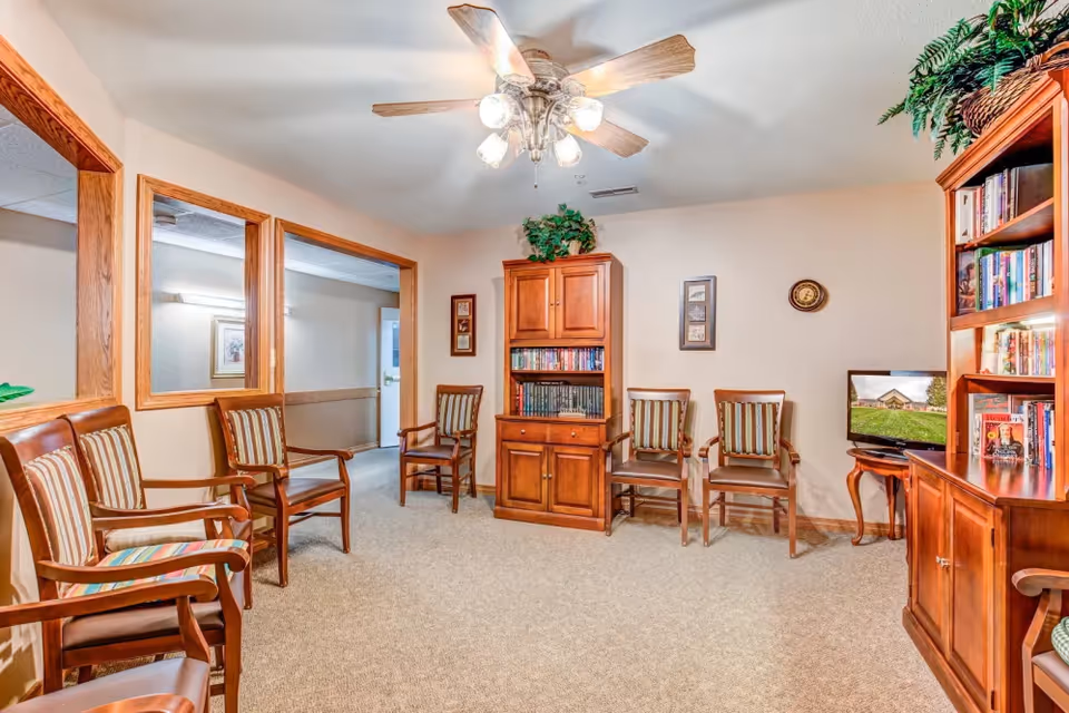 Bright common room with wooden chairs arranged around bookshelves, a small TV, and a ceiling fan.