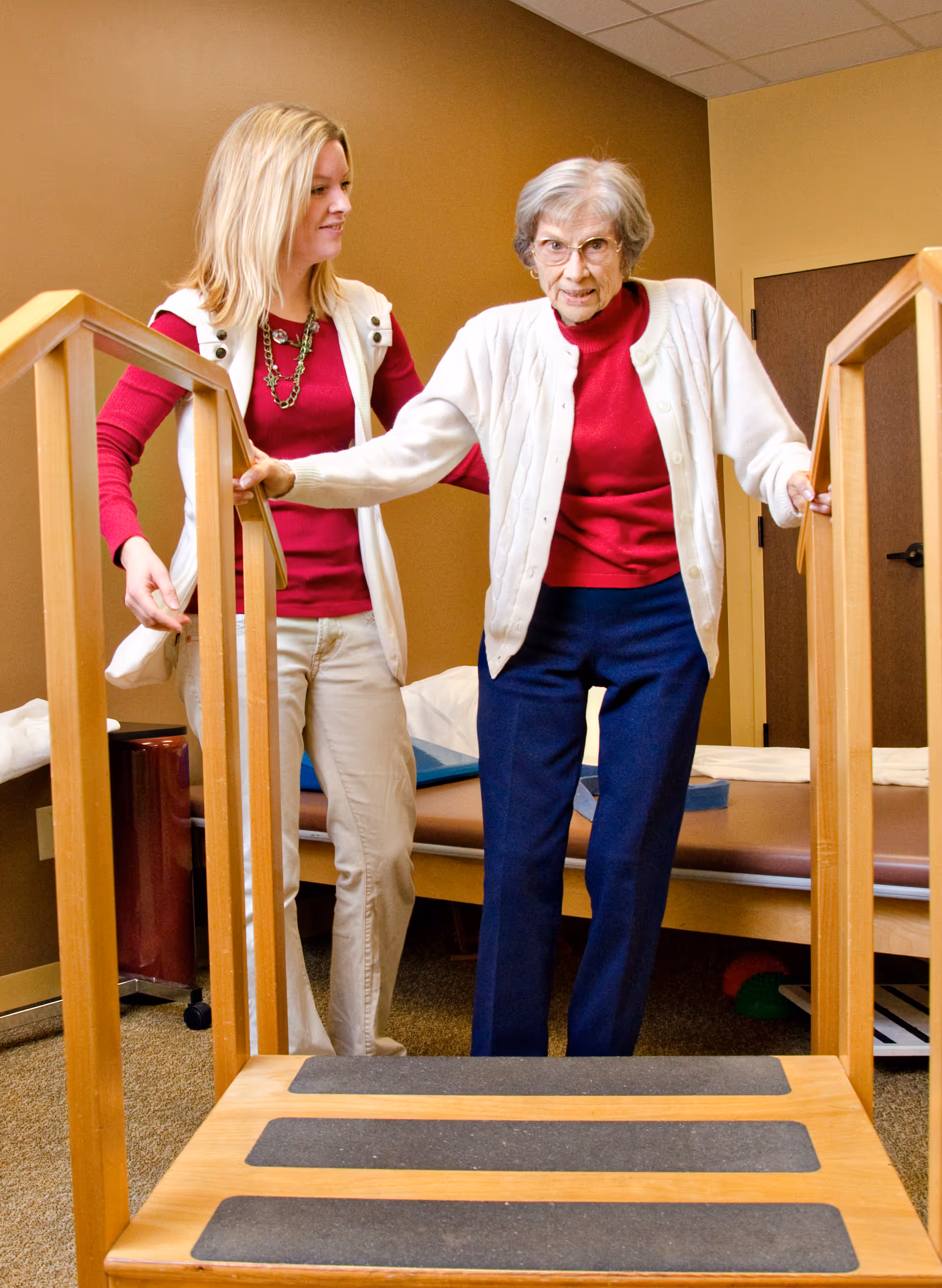 An elderly woman wearing glasses, a white cardigan, red shirt, and blue pants is holding onto wooden handrails while walking up a small set of stairs. A younger woman in a red shirt and white vest is standing beside her, providing support. They are in a room with brown walls and a treatment table in the background.