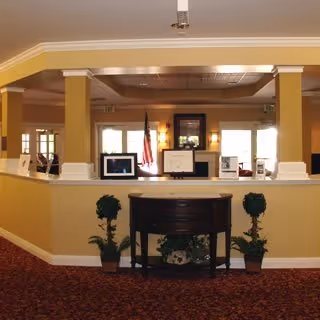 Interior view of a reception or common area with a wooden console table against a half wall, two potted plants on either side, and framed pictures and an American flag displayed on the ledge above the half wall. The space has beige walls, white trim, and a red patterned carpet.