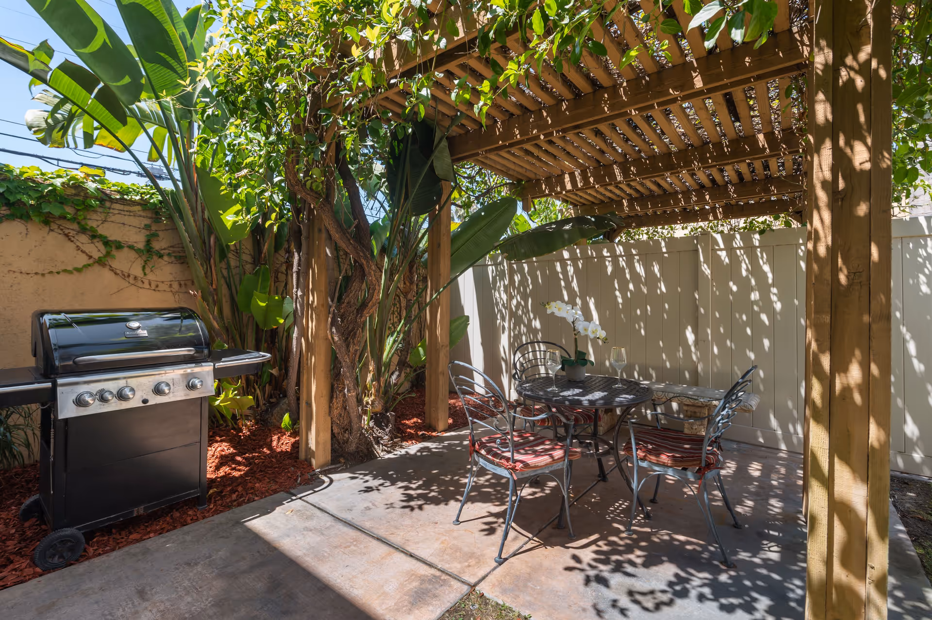Shaded backyard patio under a wooden pergola with a metal dining set, potted orchid, and a gas grill beside tropical plants.