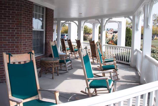 A covered porch area with several wooden rocking chairs featuring green cushions arranged along the railing. The porch has white railings and columns, with a brick wall and window on one side. In the background, there is a view of a lawn and some trees.