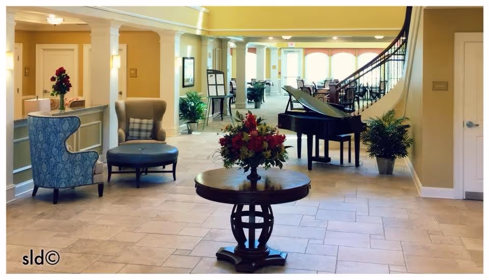 Spacious and well-lit senior living facility common area with a round wooden table holding a floral arrangement in the center. To the left, there are two upholstered chairs, one blue patterned and one beige with a plaid pillow. In the background, a black grand piano is positioned near a staircase with a decorative railing. Several potted plants and framed pictures decorate the space, which features beige walls and tiled flooring.