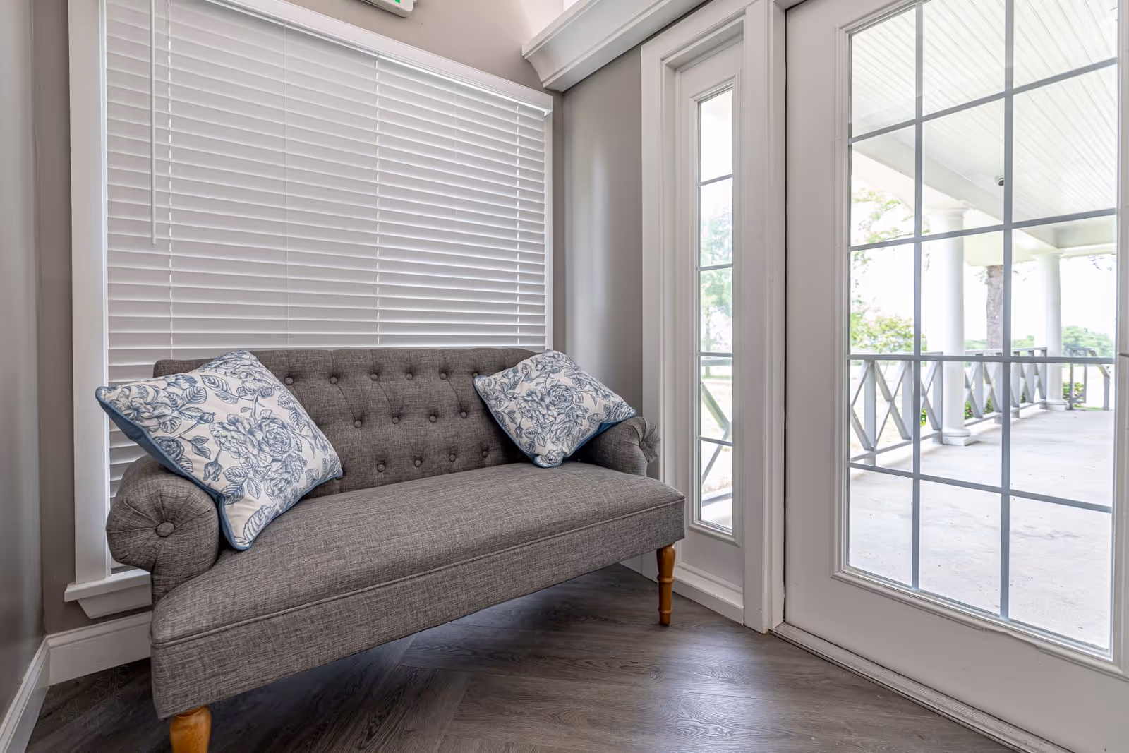 A small gray upholstered loveseat with two floral patterned pillows sits in front of a window with closed white blinds. To the right, there is a glass door with white framing leading to a covered outdoor porch area with white columns and railings.