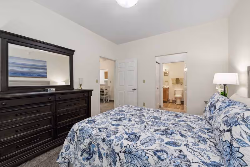 A bedroom with a large bed covered in a blue and white floral patterned bedspread. To the left is a dark wooden dresser with a large mirror. Two doorways are visible, one leading to a bathroom with a toilet and towels, and the other leading to another room with a table and chairs. The walls are painted white and the room is well-lit.