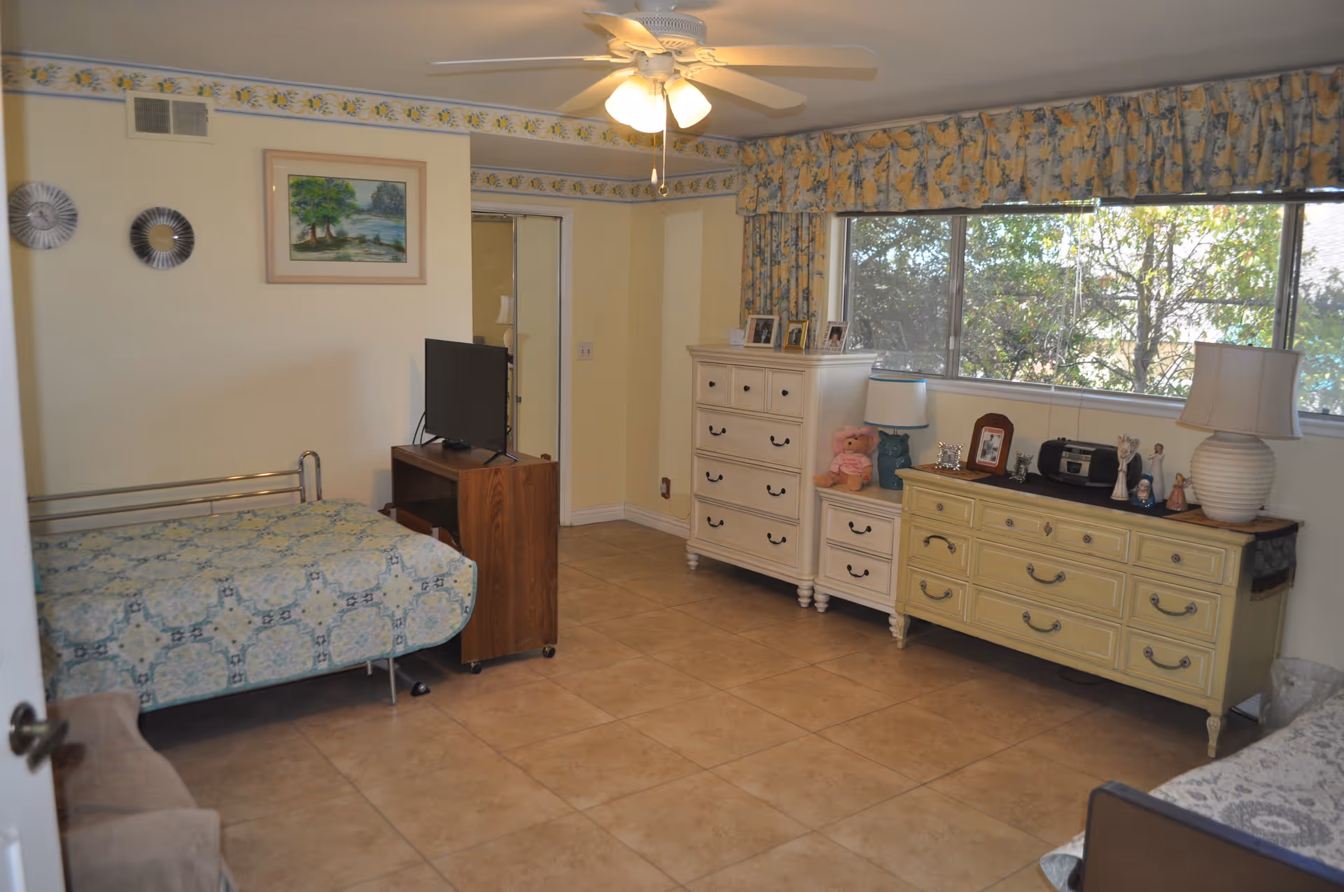 A bedroom with a single bed covered in a blue patterned quilt, a small wooden TV stand with a flat-screen TV, two dressers with various decorative items and framed photos, a large window with floral curtains, and a ceiling fan with lights.