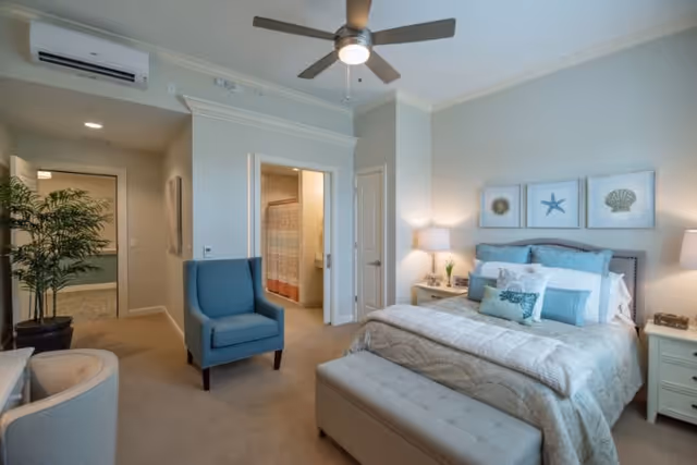 A cozy bedroom with a neatly made bed featuring multiple pillows and a cushioned bench at the foot. There is a blue armchair near the entrance to a bathroom with a shower curtain visible. The room has soft lighting from a bedside lamp and three framed sea-themed artworks above the bed. A ceiling fan is mounted on the ceiling, and a potted plant is placed near the hallway.
