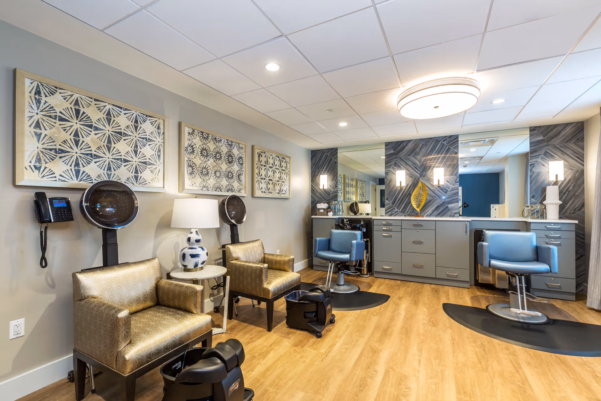 A modern salon area in a senior living facility with two blue salon chairs in front of a large mirror and gray cabinetry. To the left, there are two gold-patterned armchairs with hair dryers above them, a small round table with a decorative lamp, and three framed abstract artworks on the wall. The floor is wood, and the ceiling has recessed lighting.