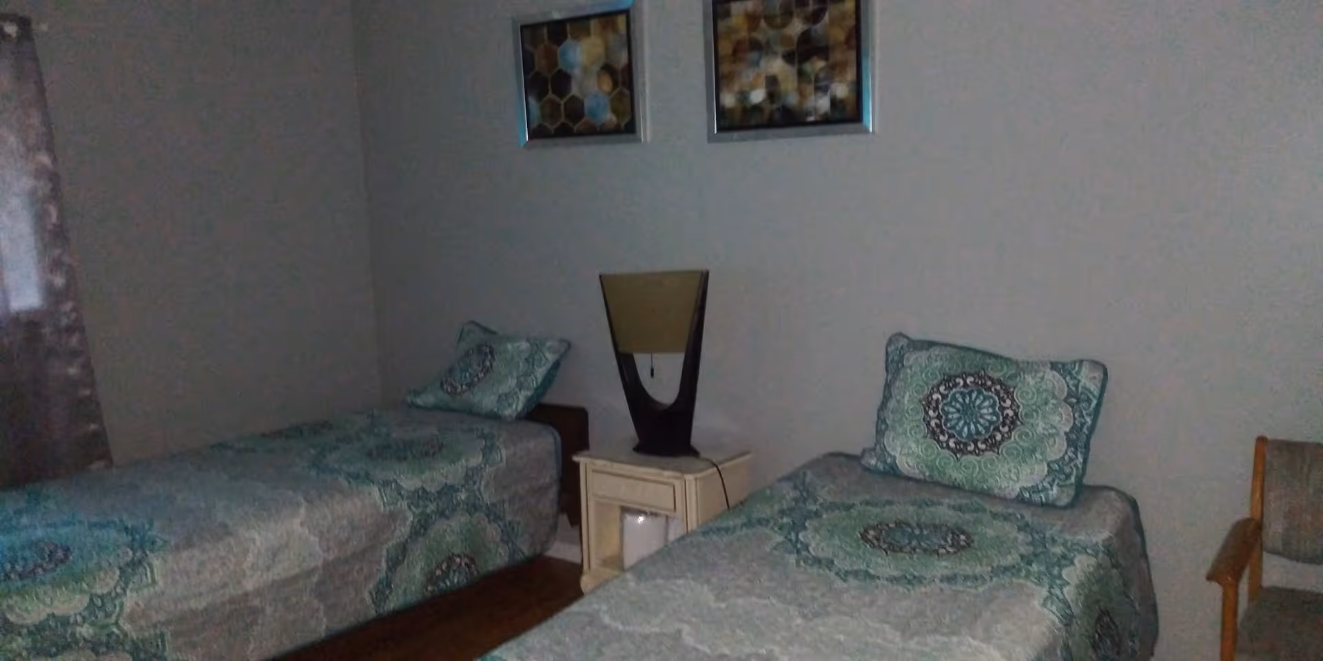 A bedroom with two single beds placed parallel to each other, each with matching green and blue patterned bedspreads and pillows. Between the beds is a small white nightstand with a modern lamp on top. There are two framed abstract artworks hanging on the wall above the nightstand. A wooden chair is positioned to the right side of the right bed. The room has a window with curtains on the left side.
