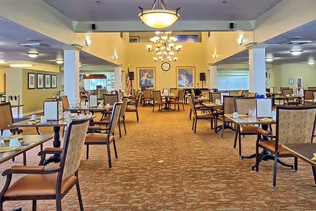 Spacious dining room with multiple tables and chairs arranged neatly. The room features warm lighting from chandeliers and ceiling lights, framed artwork on the walls, and a carpeted floor. The setting appears calm and inviting, suitable for group dining.