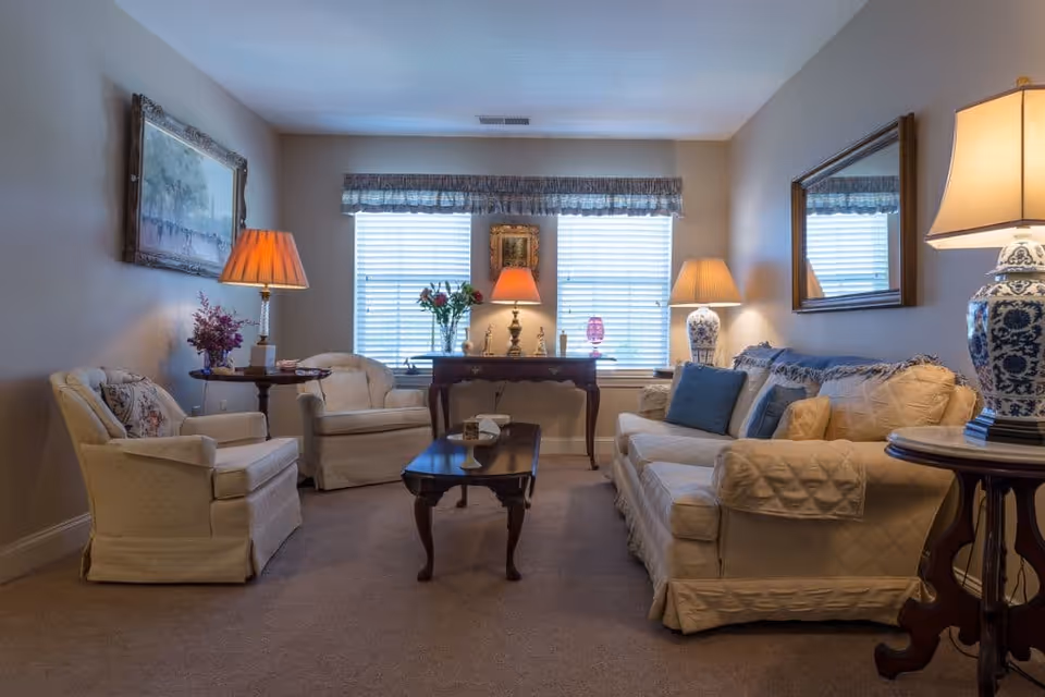 A cozy living room with beige walls and carpet, featuring a cream-colored sofa with blue and beige cushions, two matching armchairs, a wooden coffee table, and side tables with decorative lamps. There are two windows with blinds and valances, a large mirror on one wall, and framed artwork on another. The room is softly lit by multiple lamps.