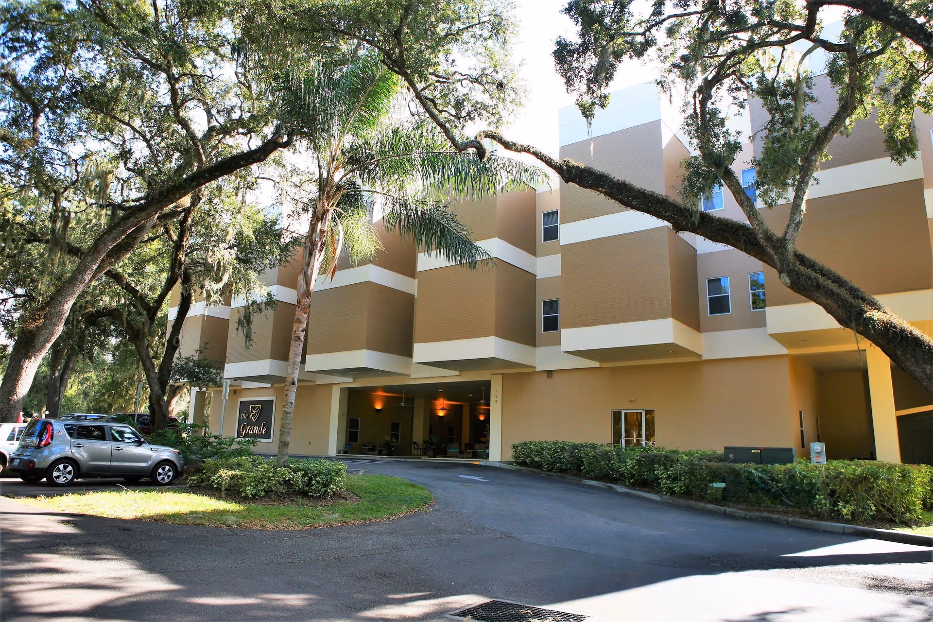 Exterior view of The Grande senior living facility showing a multi-story building with beige and white accents, surrounded by trees and parked cars in the driveway.
