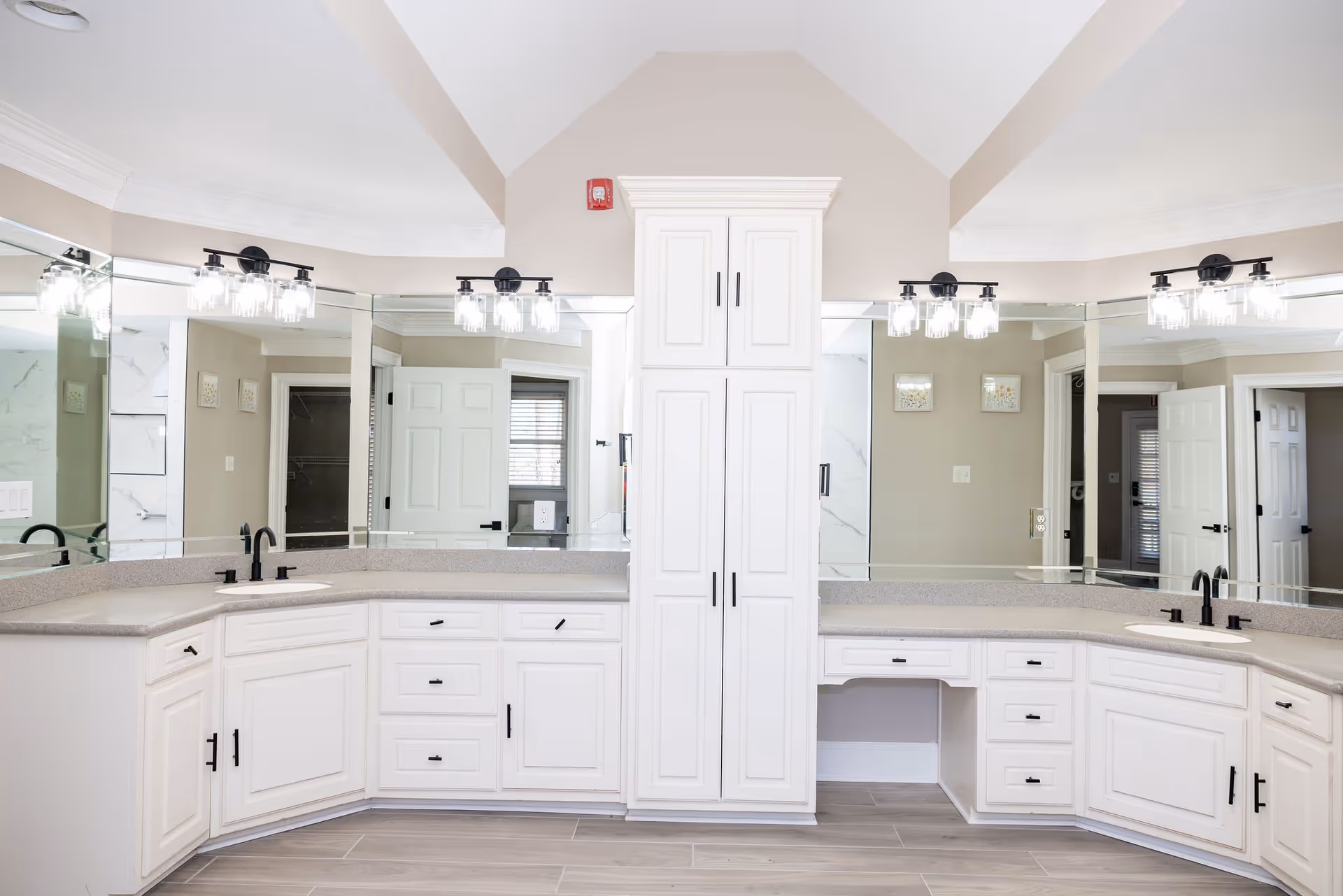Bright and spacious bathroom with dual sinks, white cabinetry, large mirrors, and modern light fixtures above each sink. The room features a central tall cabinet, beige walls, and light wood flooring.