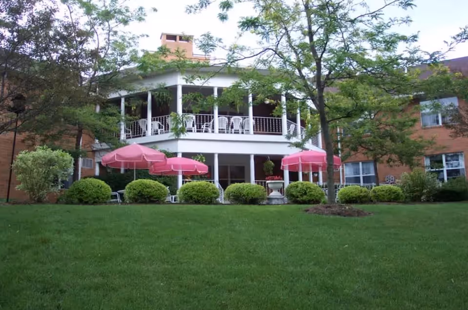 Two-story brick senior living building with a covered wraparound porch, pink patio umbrellas, manicured shrubs and a green lawn.