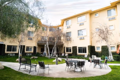 Outdoor courtyard area of a senior living facility with a circular concrete pathway, metal chairs and tables, benches, green grass, trees, and a multi-story beige building in the background.