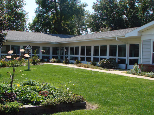 A single-story building with large windows surrounded by a well-maintained lawn and garden. There are various plants and flowers in the garden bed, and a birdhouse is visible near the building. Trees are in the background under a clear sky.