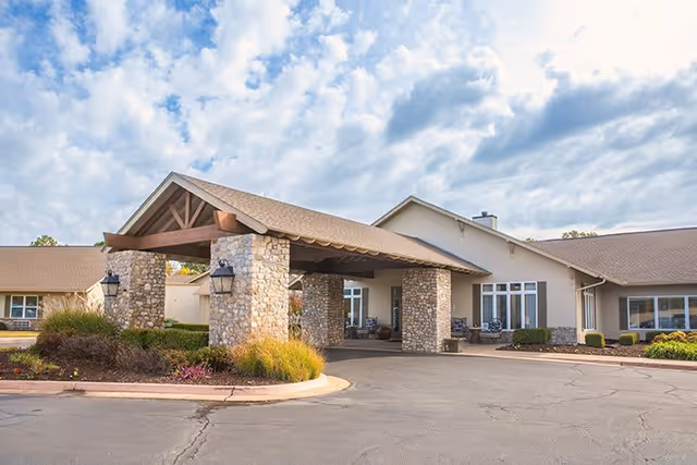Exterior view of Cedar Ridge Senior Living facility showing a covered entrance with stone pillars, beige walls, and a paved driveway under a partly cloudy sky.