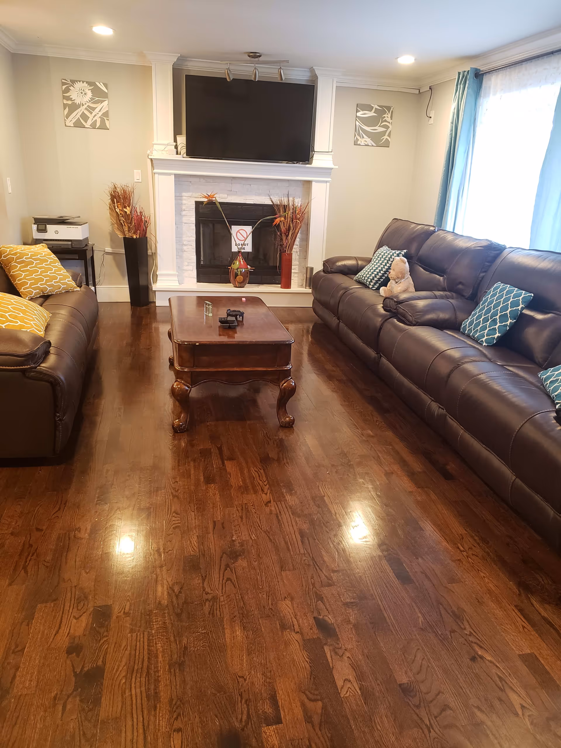 Bright living room with dark hardwood floors, two brown leather sofas, a central wooden coffee table, and a fireplace with a TV mounted above.