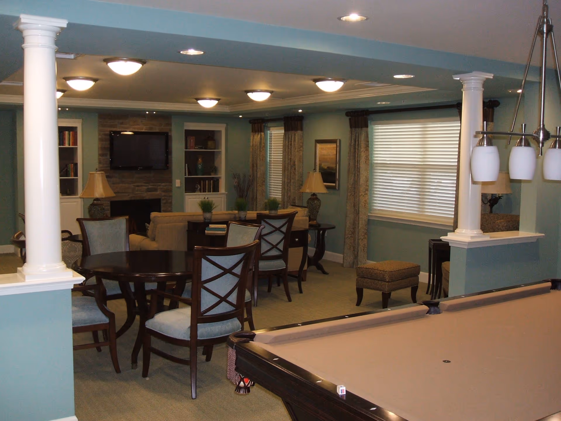 Common area with a pool table in the foreground, round table and chairs, a seating area with a TV above a fireplace, and decorative columns.