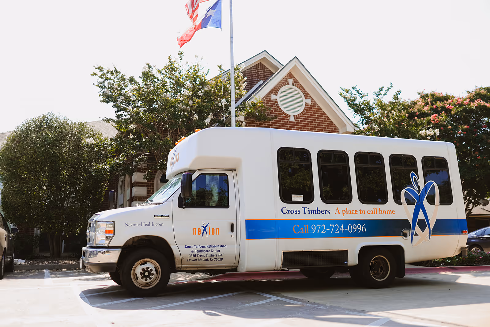White Cross Timbers shuttle bus parked outside a brick rehabilitation and healthcare center under trees and flags.