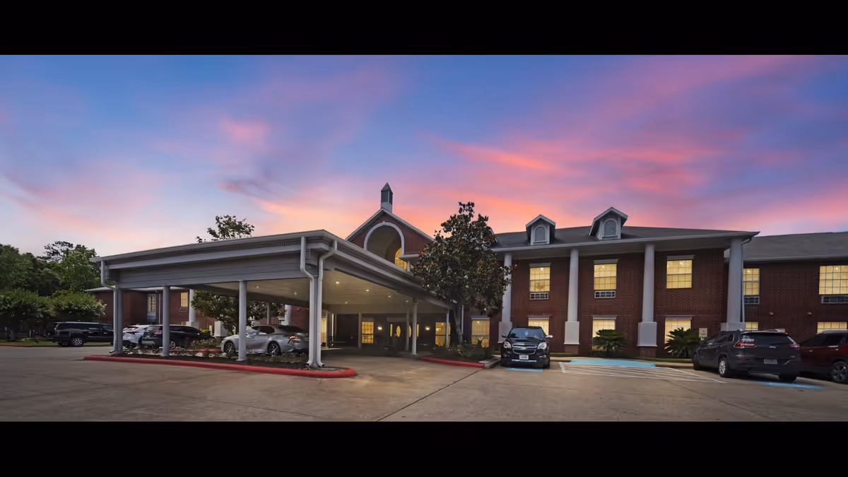 Exterior view of Collier Park facility at sunset with a covered entrance, parked cars, and a two-story brick building with white columns and lit windows.