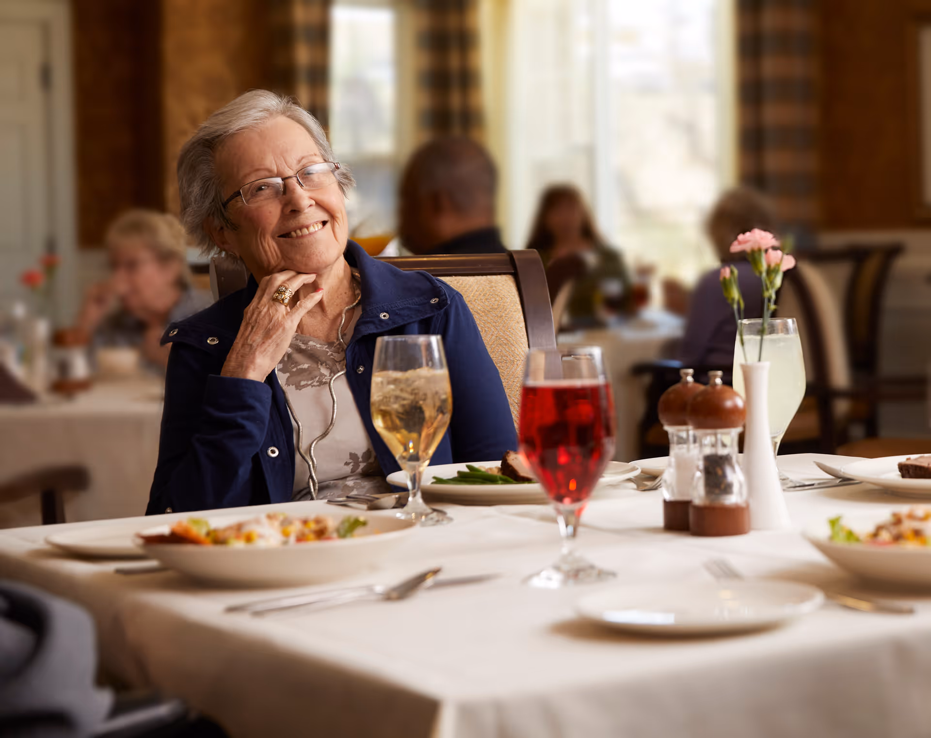 An elderly woman with glasses and a navy blue jacket smiling while sitting at a dining table in a senior living facility. The table is set with plates of food, glasses of beverages, salt and pepper shakers, and a small vase with pink flowers. Other people are seated and dining in the background.