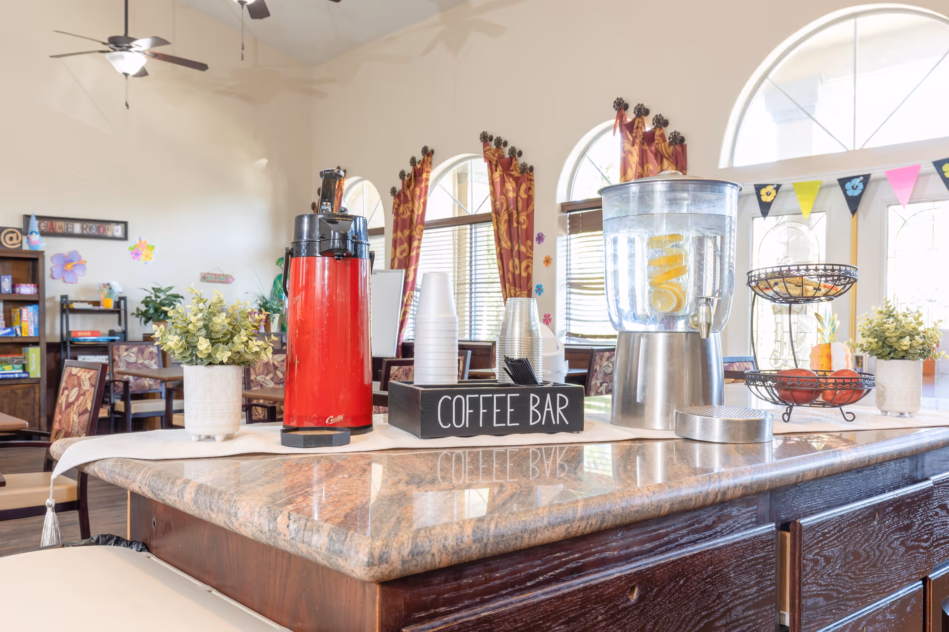 A bright and inviting coffee bar area in a senior living facility with a red coffee dispenser, a water dispenser with lemon slices, disposable cups, and a two-tiered fruit basket on a marble countertop. The background shows a dining area with tables, chairs, decorative curtains, and large arched windows letting in natural light.