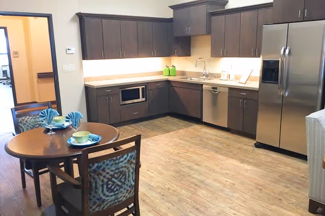 A modern kitchen area with dark wood cabinets, stainless steel refrigerator and dishwasher, a microwave built into the lower cabinets, and a round wooden dining table set with two chairs. The table is set with blue napkins and green bowls. The floor is wood-style laminate, and the walls are light-colored.