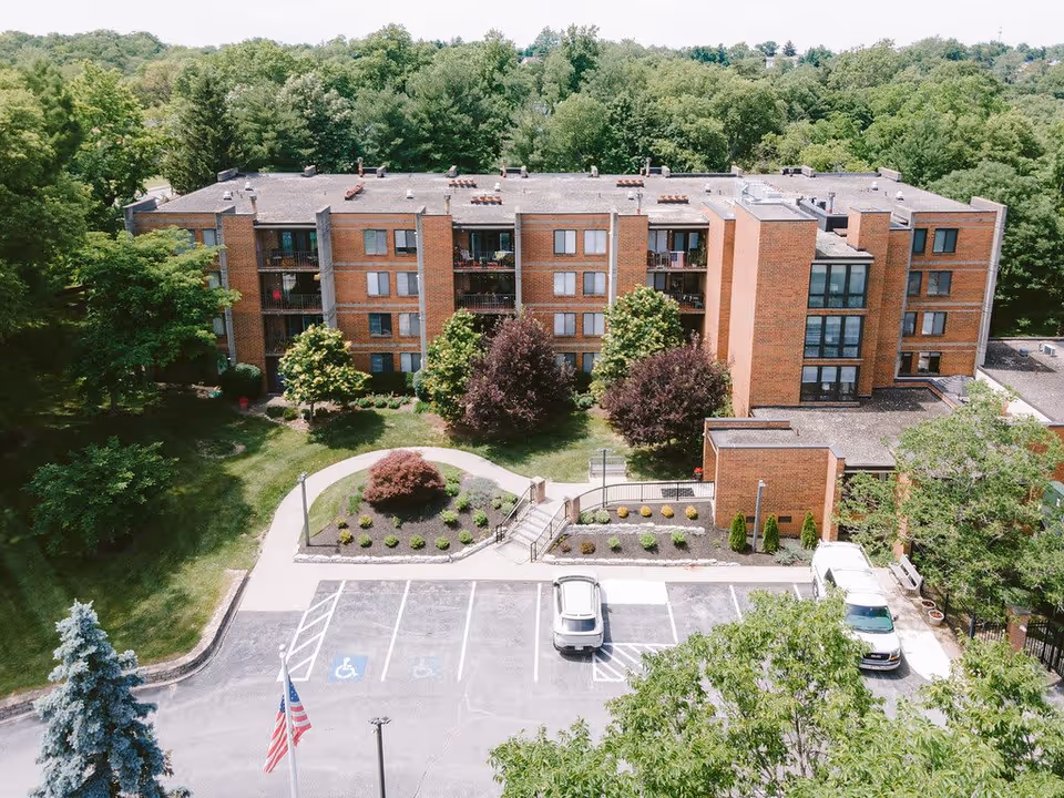 Aerial view of a multi-story brick building surrounded by trees and greenery. The building has balconies and multiple windows. In front of the building is a small landscaped area with bushes and a pathway leading to the entrance. There is a parking lot with a few cars and handicap parking spaces, and an American flag is visible near the parking area.