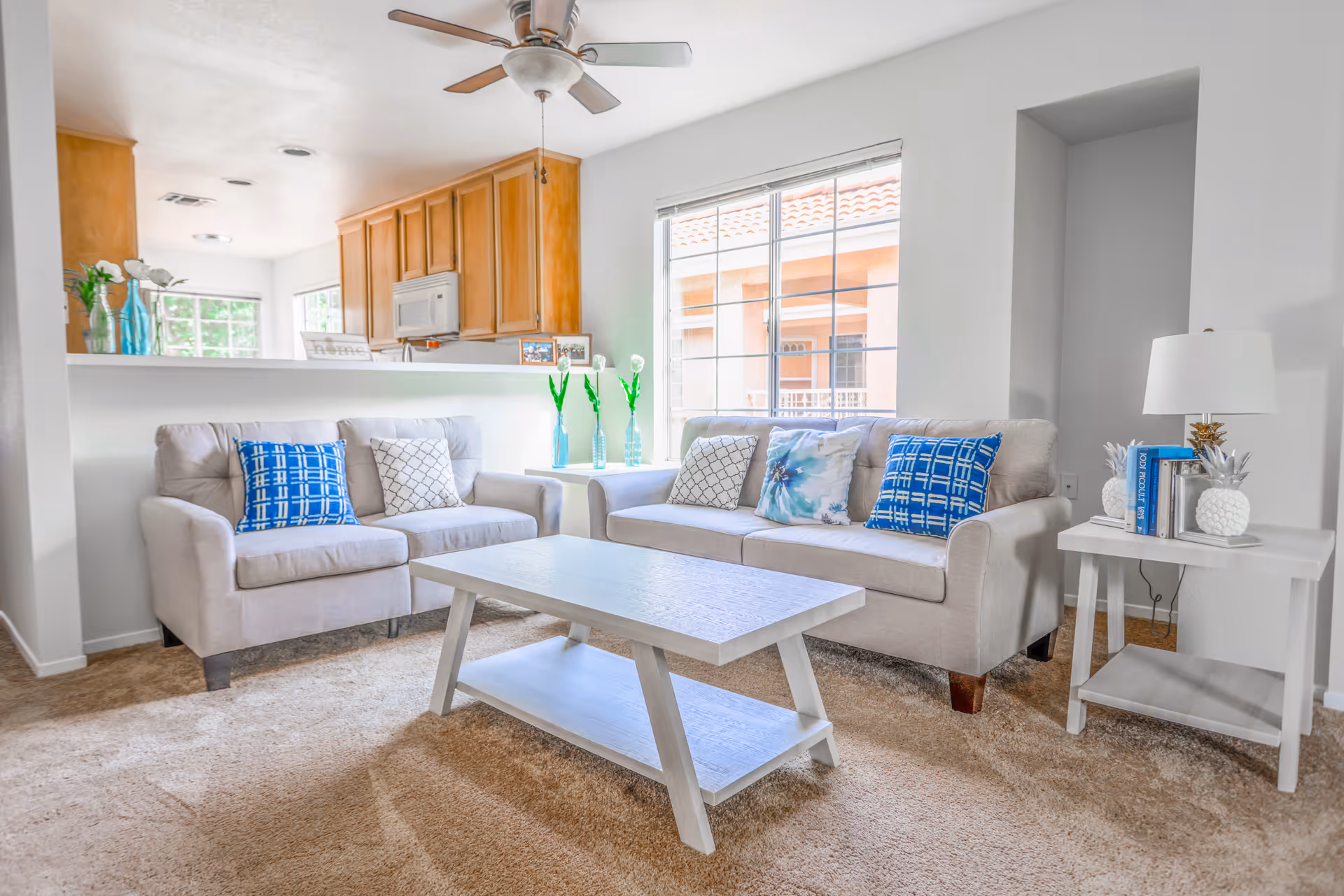 A bright living room with two beige sofas adorned with blue and patterned throw pillows, a white wooden coffee table, and a side table with a lamp and decorative items. The room has a large window letting in natural light and an open view into a kitchen with wooden cabinets and a ceiling fan above.