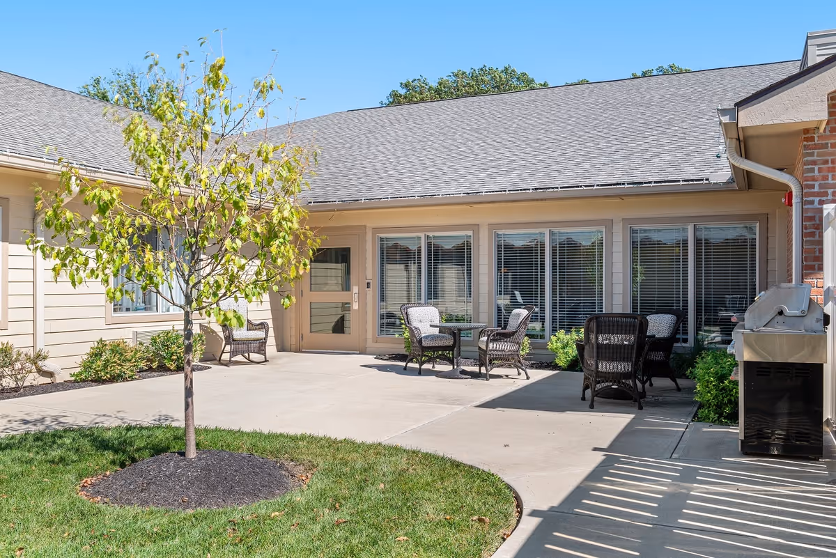 Outdoor patio area at Sage Park Assisted Living & Memory Care with a small tree planted in a mulch bed surrounded by grass, several wicker chairs with cushions arranged around small tables, and a barbecue grill on a concrete patio next to a building with large windows and a door.